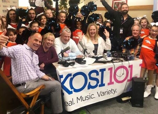 KOSI 101 staff at a table surrounded by young girls wearing Denver Bronco Cheerleader outfits